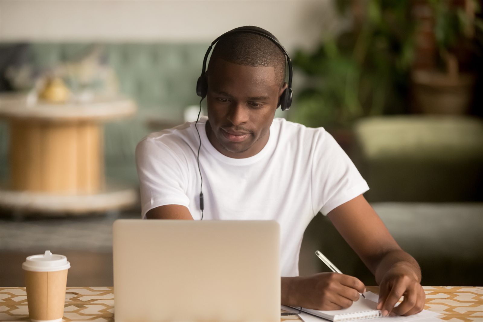 Student with headphones writing notes beside a laptop